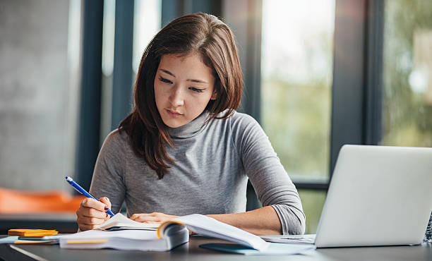 lady-at-table-working-on-laptop-while-writing-in-notebook-with-textbook-open-in-front-of-her