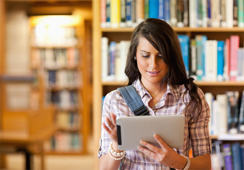 student-in-lbrary-referring-to-ipad-while-standing-with-backpack