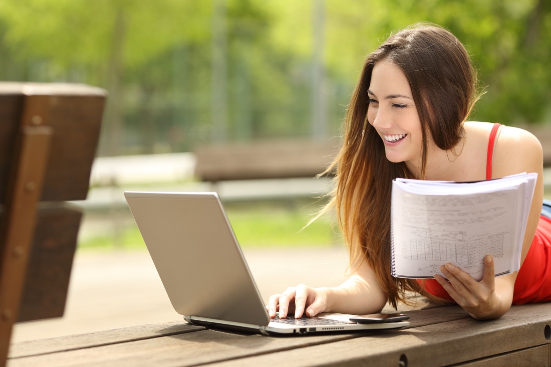 girl-outside-referring-to-notebook-while-typing-on-laptop-during-a-sunny-day