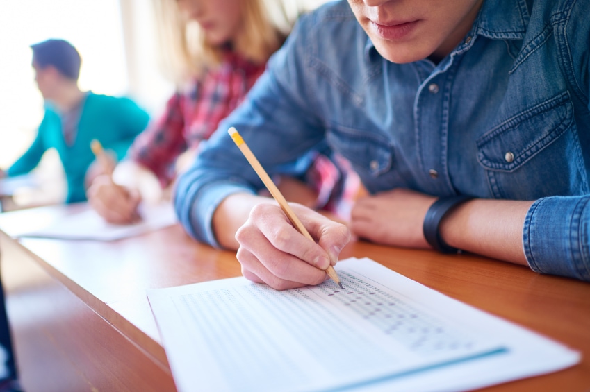 student-filling-out-mcq-answer-sheet-at-exam-with-pencil-wearing-a-denim-shirt-sitting-next-to-peers